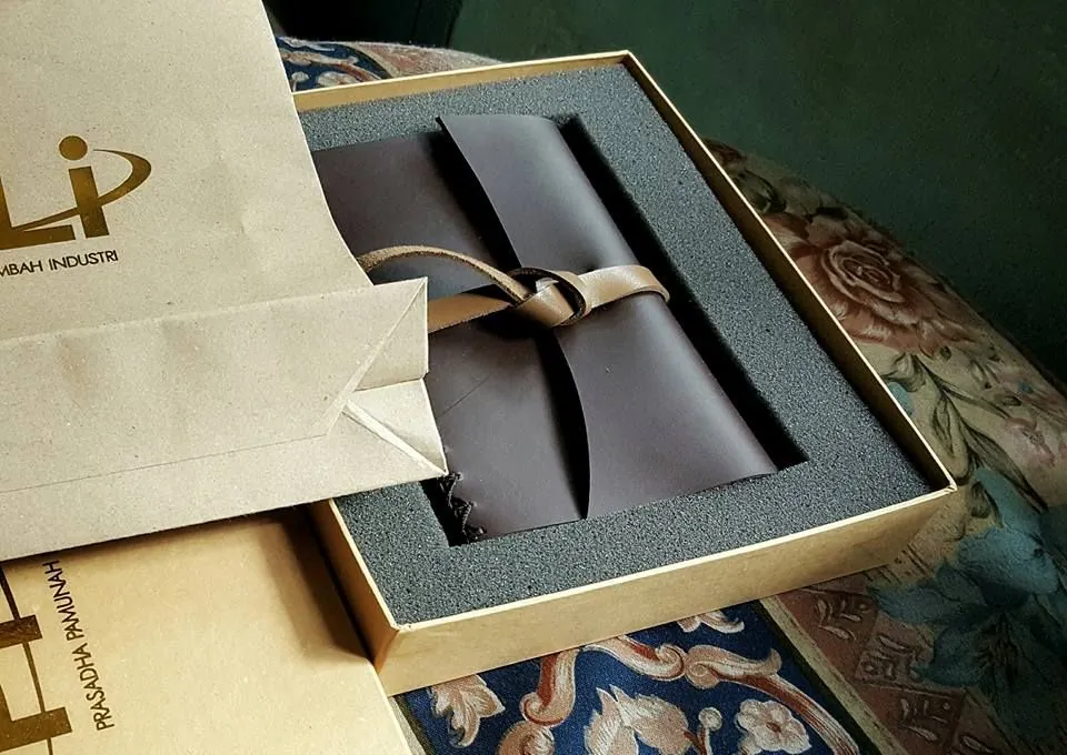 Jurnal kulit gelap diikat tali kulit, tersimpan dalam kotak hadiah berlapis busa di samping tas kertas berlogo Hibrkraft