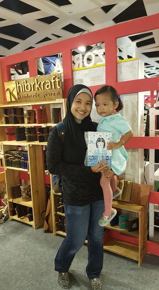 Seorang ibu berhijab menggendong anaknya yang memegang jurnal bersampul ilustrasi, berdiri di depan rak jurnal kulit Hibrkraft