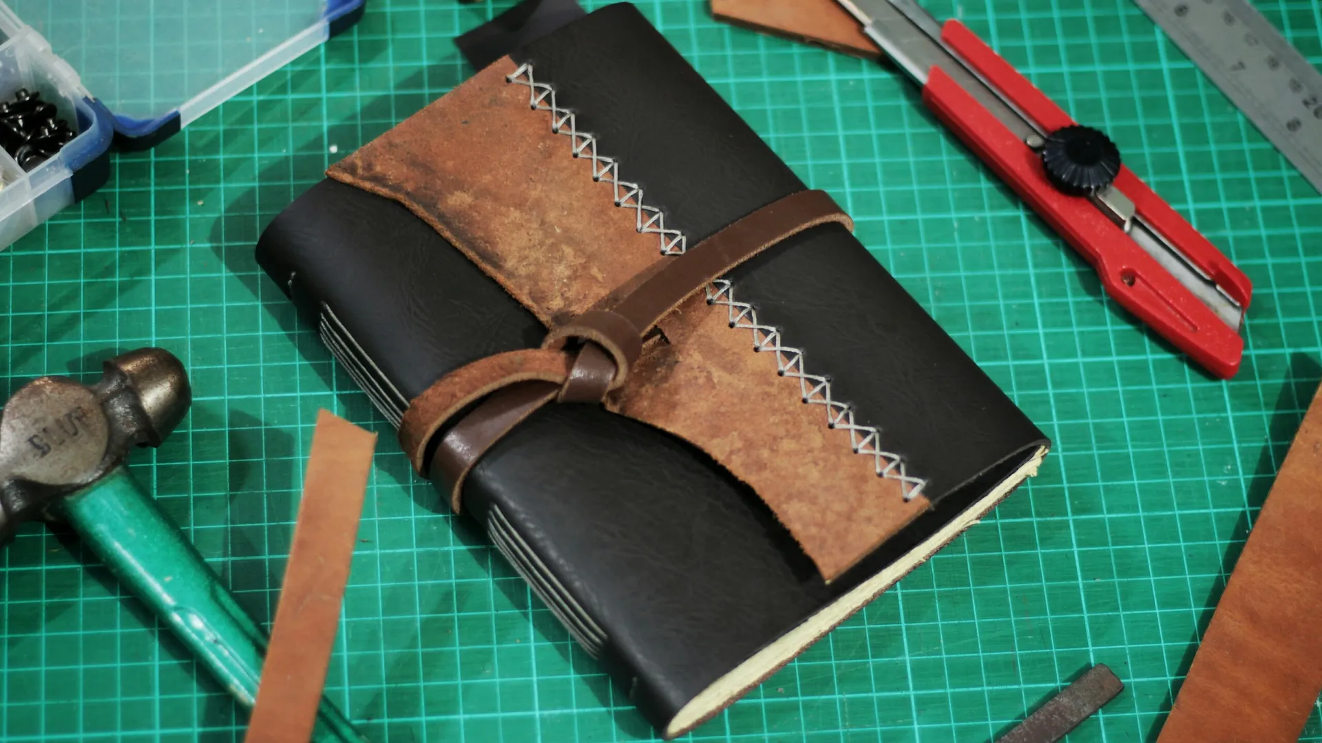 Sebuah jurnal kulit hitam dengan jahitan tangan putih sedang dibuat di atas meja potong hijau, menunjukkan proses craftsmanship.