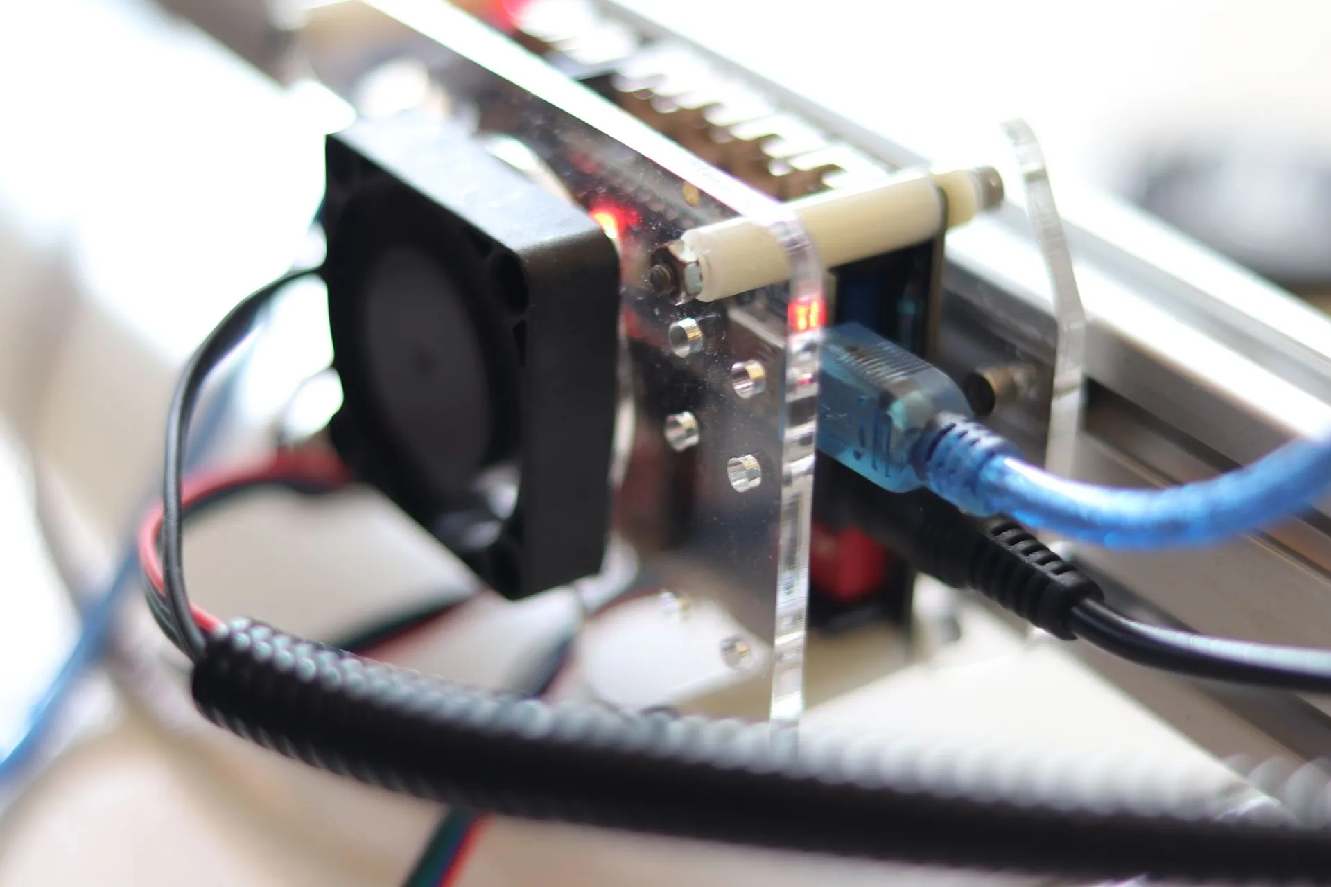 Close-up komponen elektronik dari printer 3D atau laser engraver, dengan kabel biru dan kipas pendingin hitam.