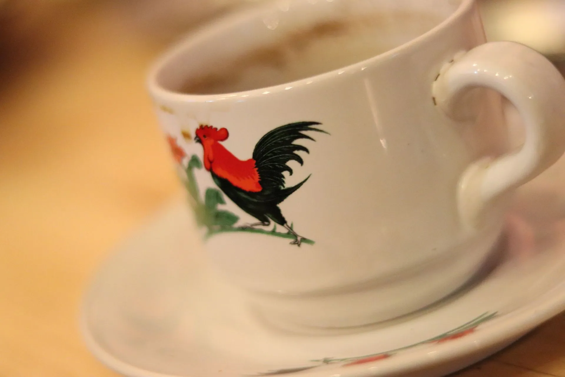 Close-up cangkir kopi putih dengan gambar ayam jago merah-hitam yang ikonik, simbol kerja keras dan budaya Indonesia.