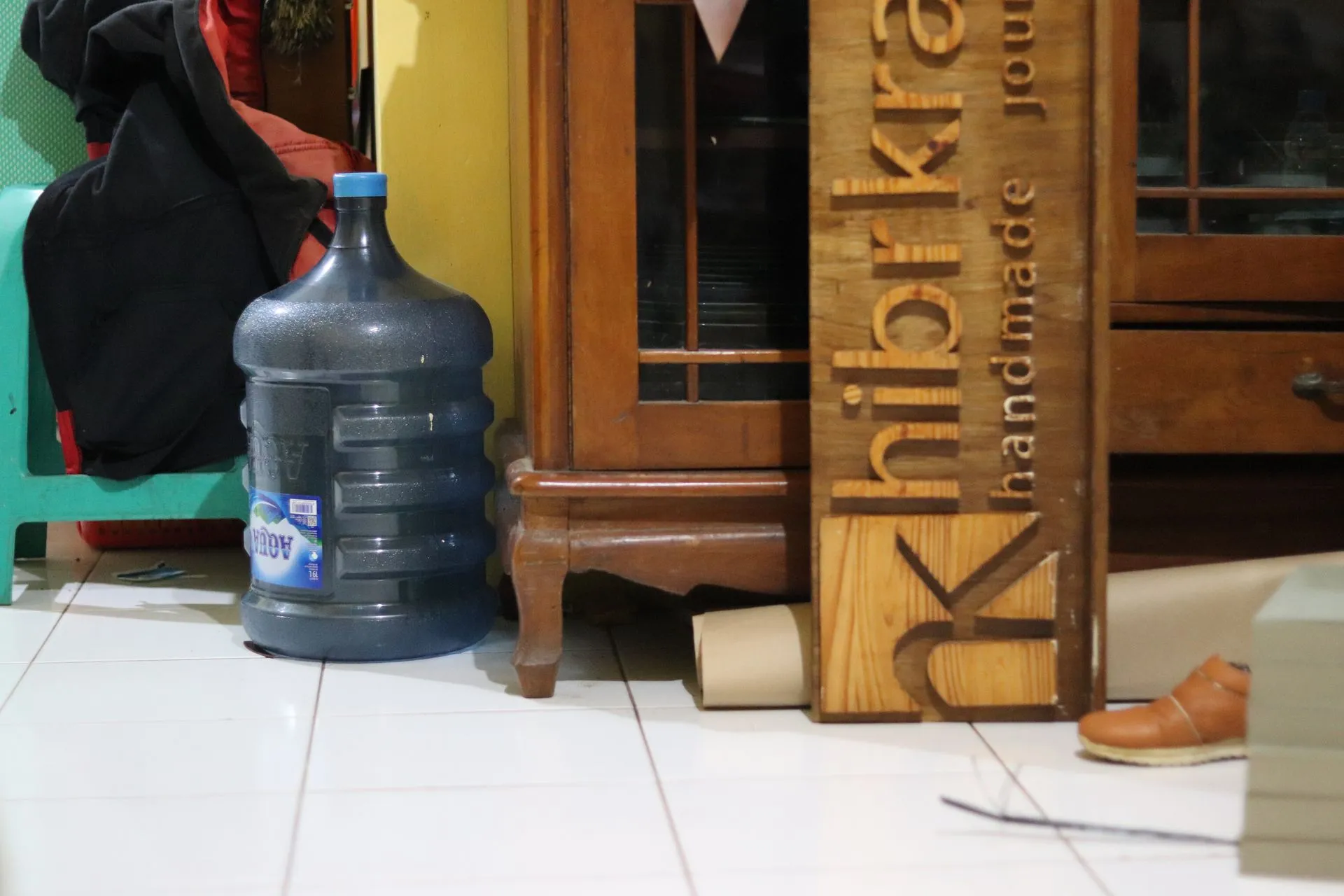 Sebuah galon air minum Aqua diletakkan di lantai sebelah papan nama Hibrkraft yang terbuat dari kayu ukir.