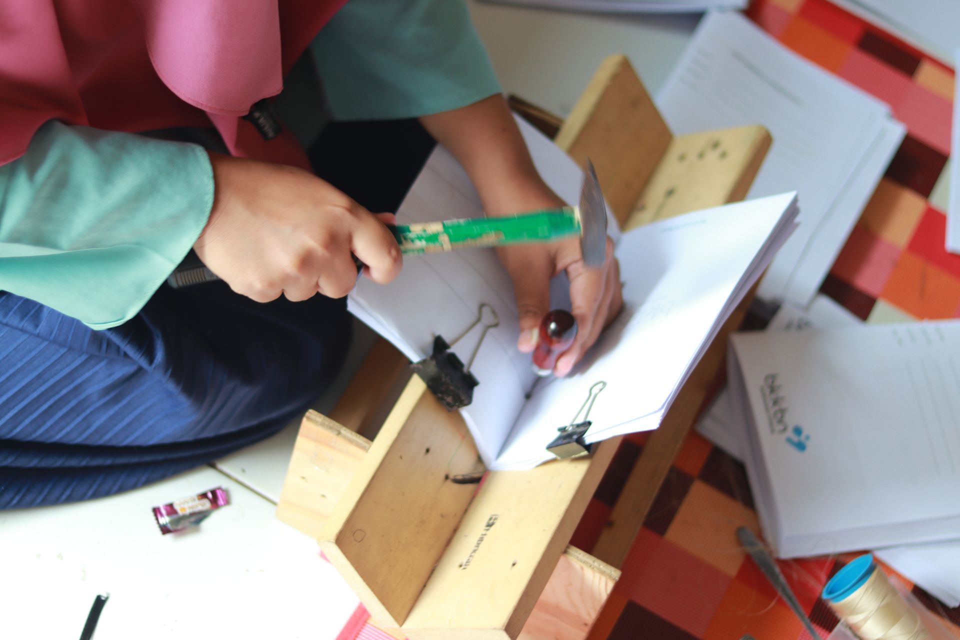 Seorang pengrajin wanita menggunakan palu dan pelubang untuk membuat lubang pada kertas yang diletakkan di dalam bookbinding cradle kayu buatan sendiri.