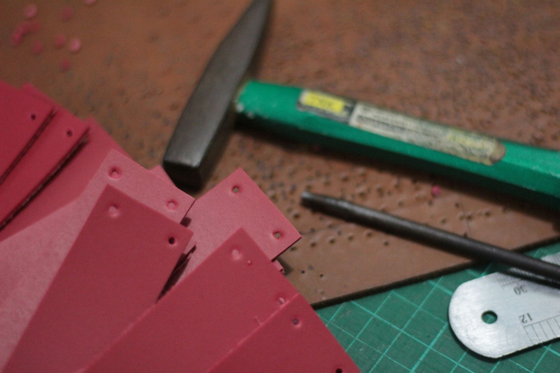 Tumpukan potongan kulit berwarna merah yang telah dilubangi dengan presisi, di sampingnya terdapat palu, pelubang, dan pemandu logam di atas cutting mat.