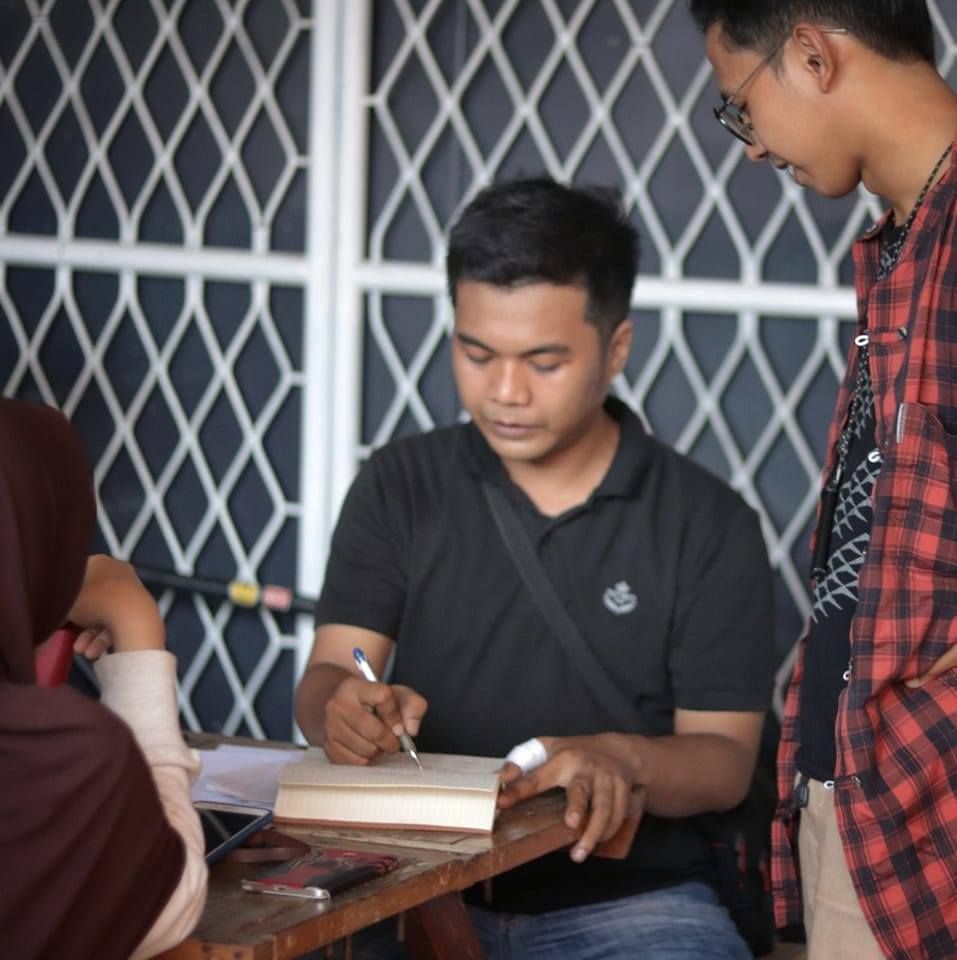 Seorang pria sedang fokus menulis atau menandatangani sebuah buku tebal di atas meja kayu.