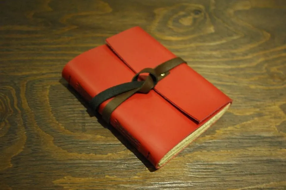 Sebuah jurnal kulit Hibrkraft Basic berwarna merah cerah tergeletak di atas meja kayu yang artistik.