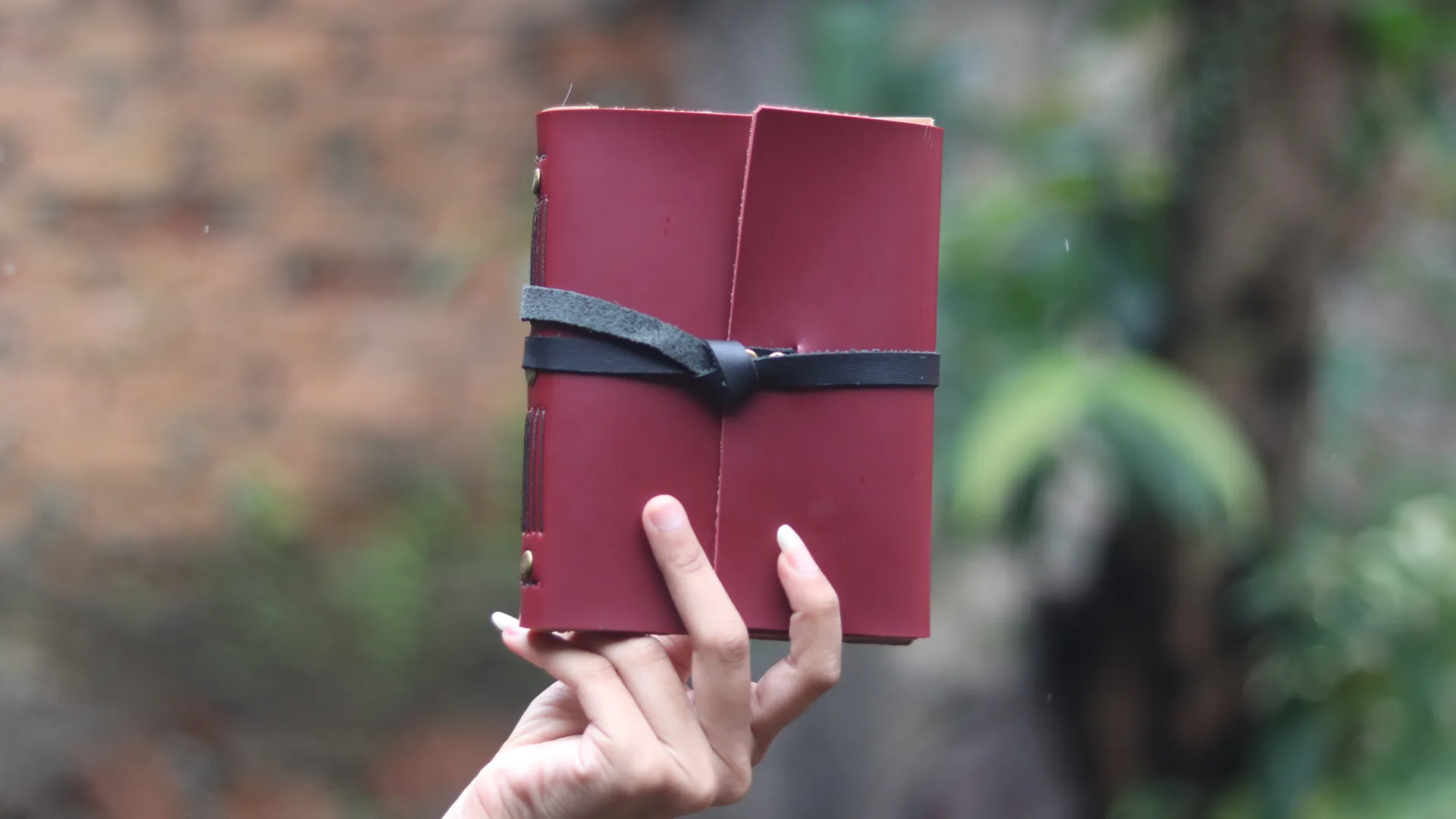 Sebuah tangan wanita dengan kuku terawat memegang agenda kulit eksklusif berwarna maroon yang elegan.