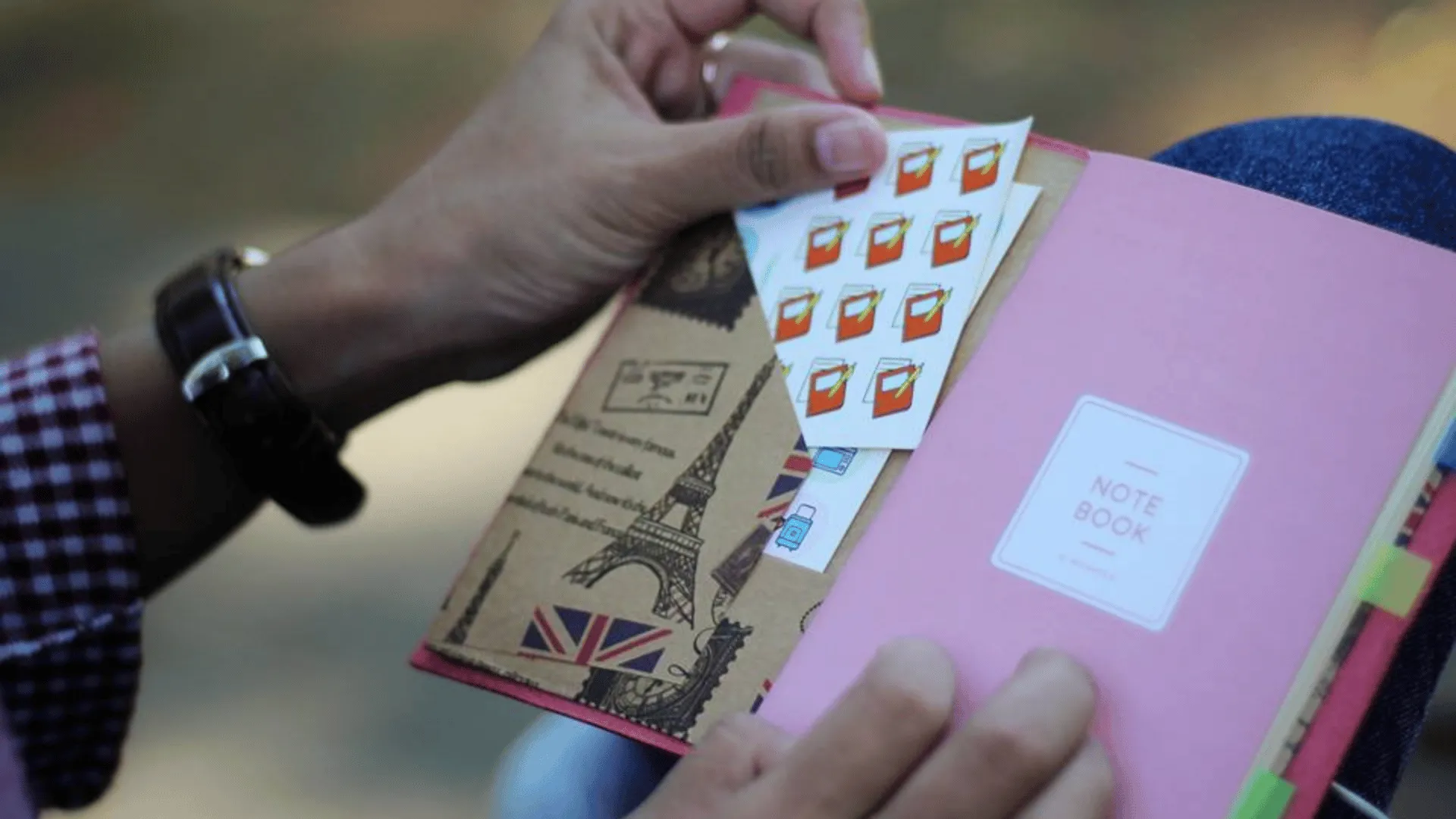 Sebuah custom notebook berwarna pink yang dihiasi stiker bertema perjalanan, menunjukkan bagaimana elemen dekoratif sederhana dapat menanamkan identitas dan semangat petualangan pada benda sehari-hari.
