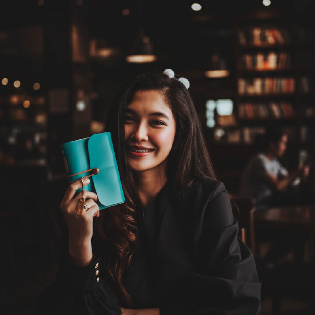 Seorang wanita tersenyum di sebuah kafe sambil memegang jurnal berwarna teal, melambangkan awal yang baru dan positif.