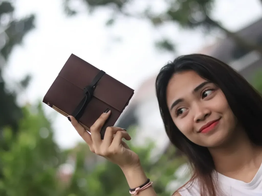 Wanita di luar ruangan tersenyum sambil memegang buku catatan kulit cokelat tua bertutup tali kulit.