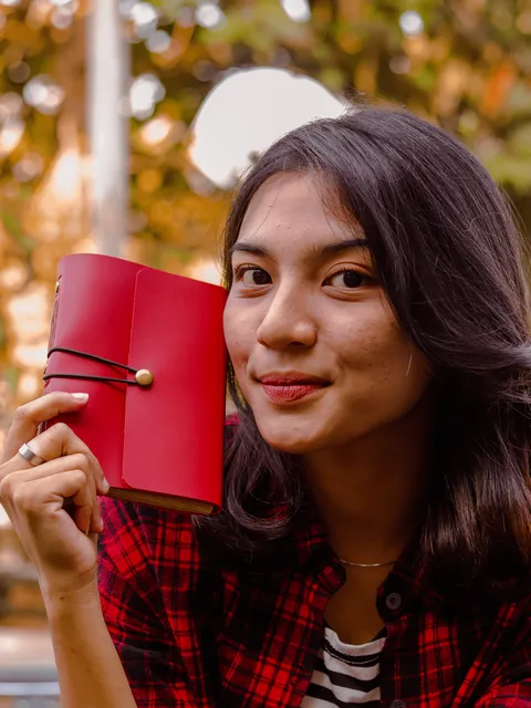 Seorang wanita dengan kemeja flanel merah tersenyum sambil memegang jurnal kulit merah cerah, melambangkan awal yang baru dan semangat.