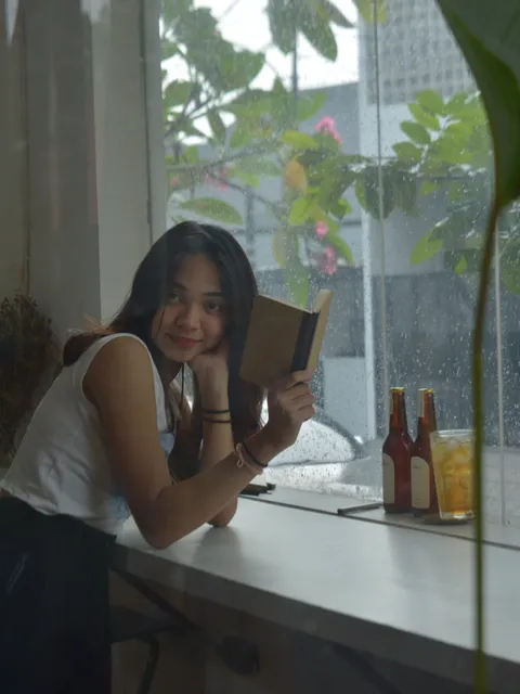 Wanita menikmati suasana hujan sambil membaca buku catatan umum, menunjukkan ketenangan dan refleksi yang bisa didapatkan.