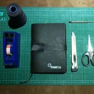 Notebook bersampul gelap di meja kerja, dikelilingi gulungan benang jahit, alat pelubang, dan gunting untuk penjilidan