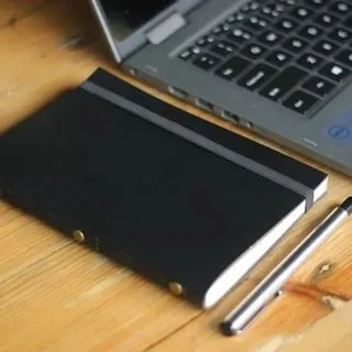 Notebook bersampul gelap di atas meja kayu, berdampingan dengan laptop dan dua pena