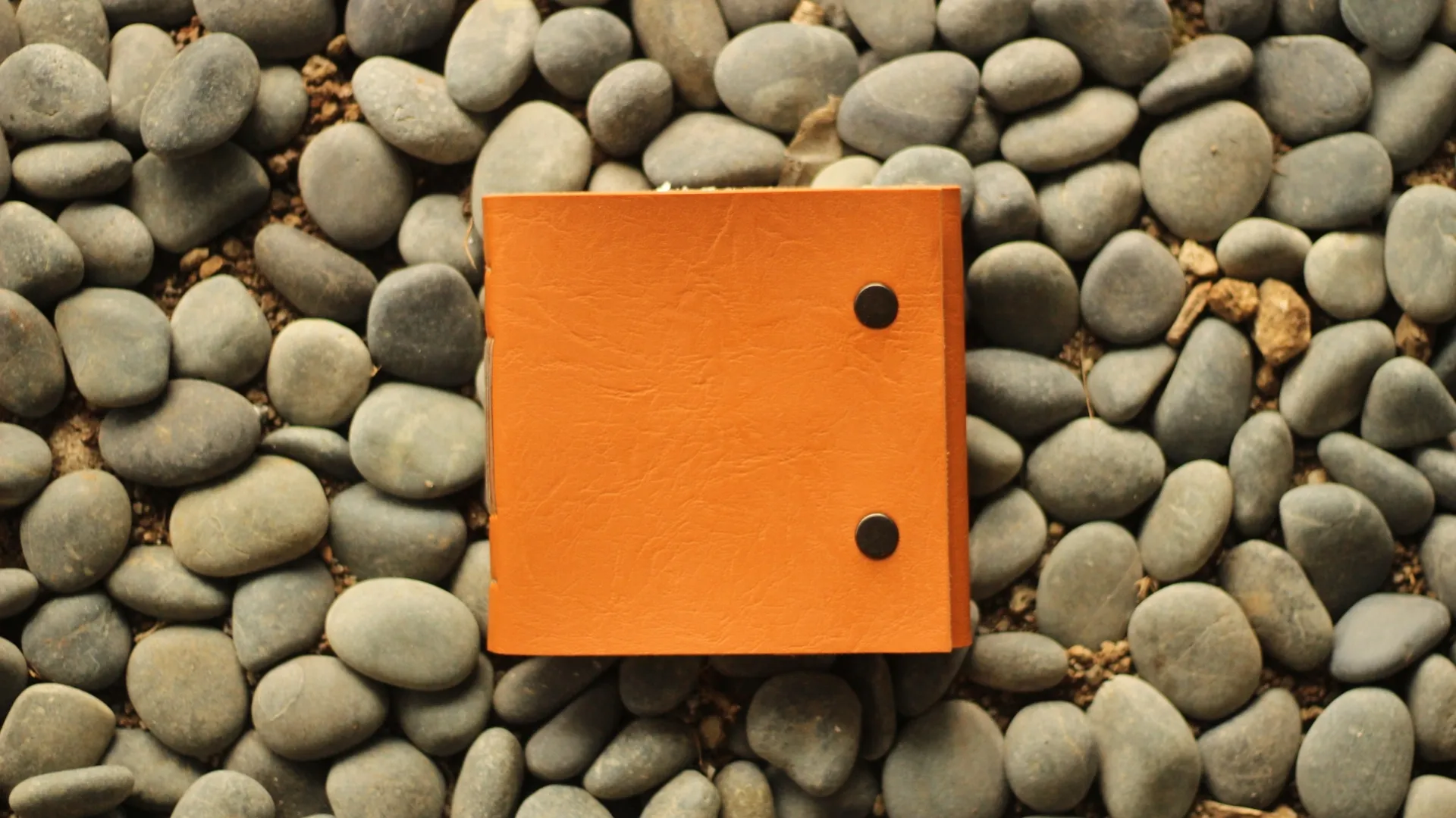 Jurnal kulit berwarna oranye diletakkan di atas bebatuan, menunjukkan daya tahan dan estetikanya yang cocok untuk arsitek.