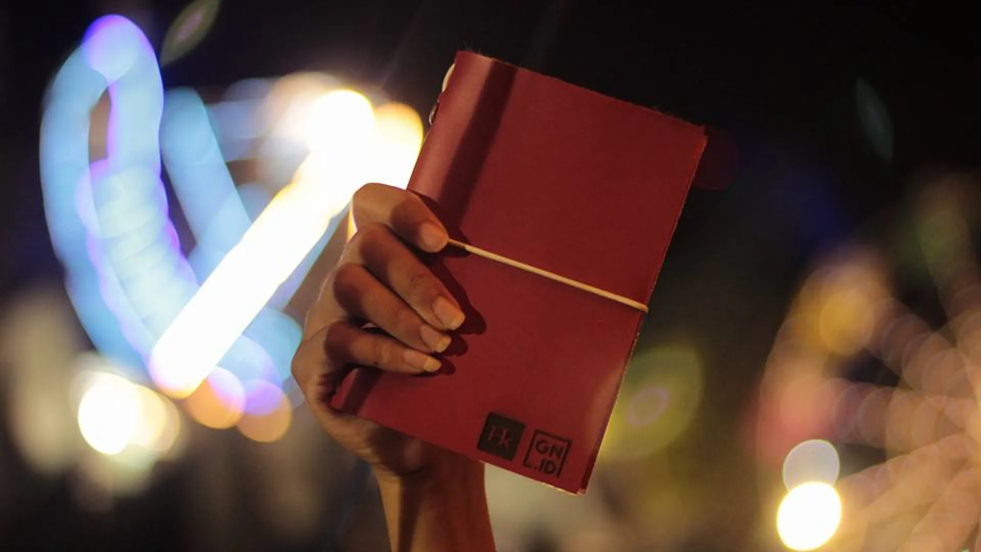 Tangan memegang jurnal kulit merah di malam hari dengan latar belakang lampu kota, menunjukkan kesiapan untuk sketching kapan saja.