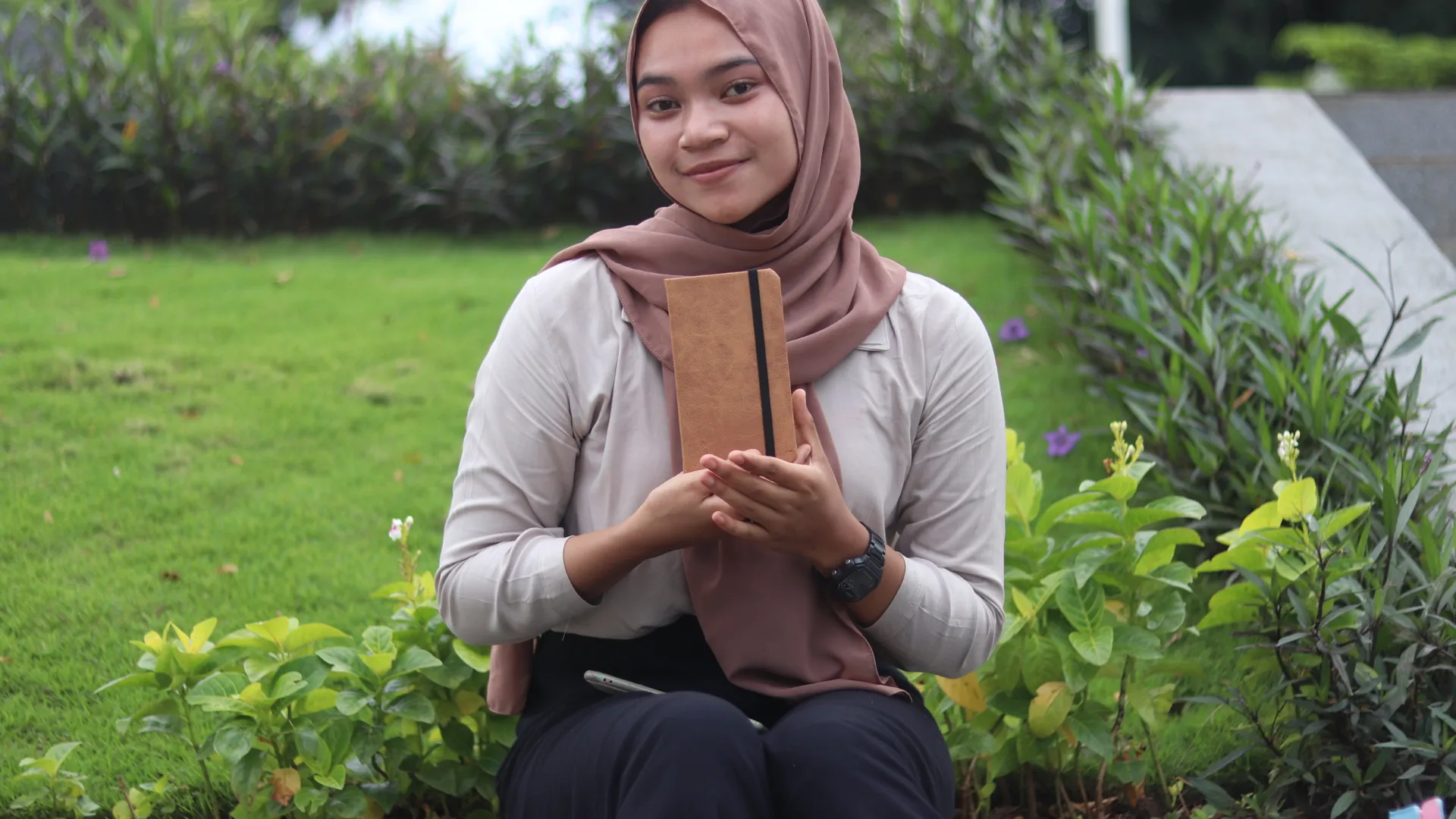 Seorang wanita memegang jurnal custom coklat di taman, mencari inspirasi untuk novelnya.