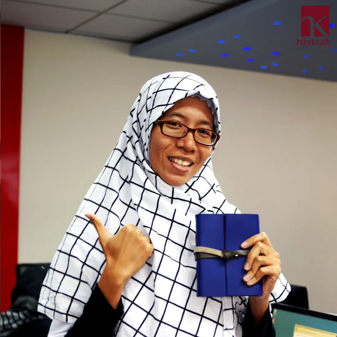 A woman in a black and white grid-patterned hijab giving a thumbs up while holding a deep blue handmade leather notebook
