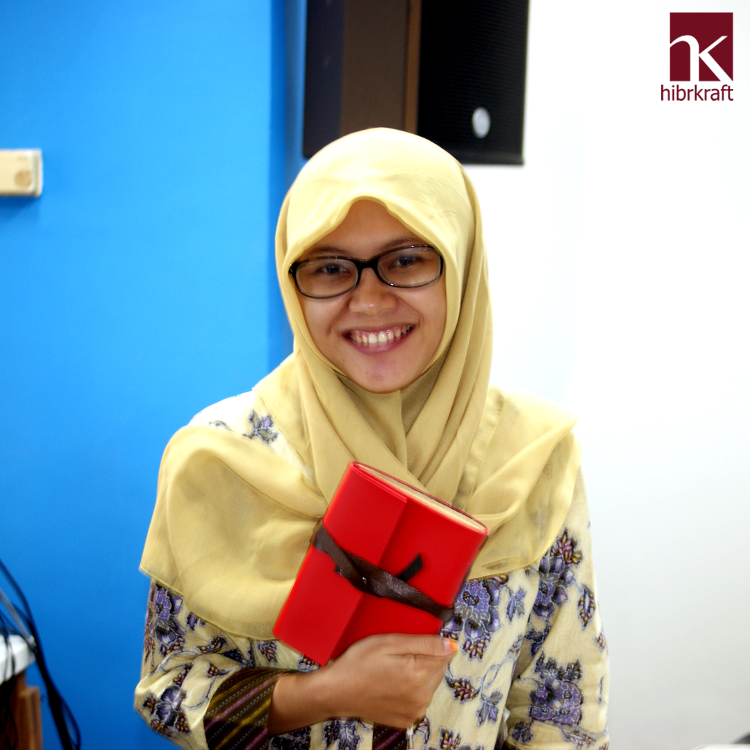 A smiling woman in a yellow hijab holding a bright red Hibrkraft custom leather journal