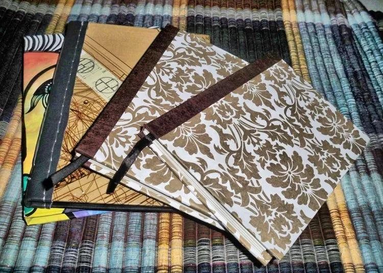 Several bound journals fanned out, a tan damask-print cover in front, with an orange illustrated cover and a cream cover carrying a technical line-drawing motif visible behind it