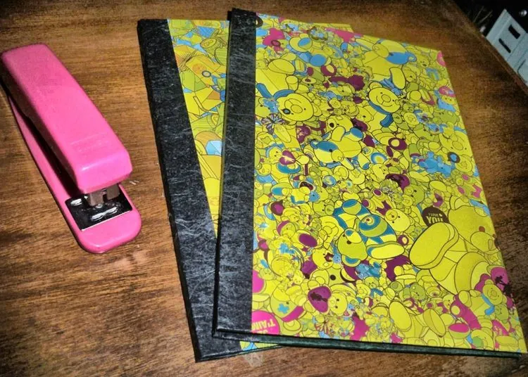 Two notebooks with an all-over yellow cartoon-bear print cover and black cloth spines, shown beside a pink stapler for scale