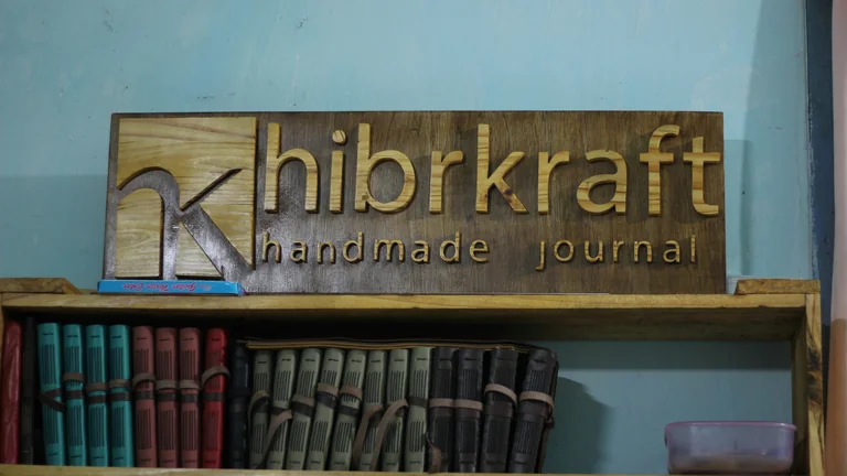 Papan kayu bertuliskan hibrkraft di atas rak berisi jurnal kulit berwarna-warni