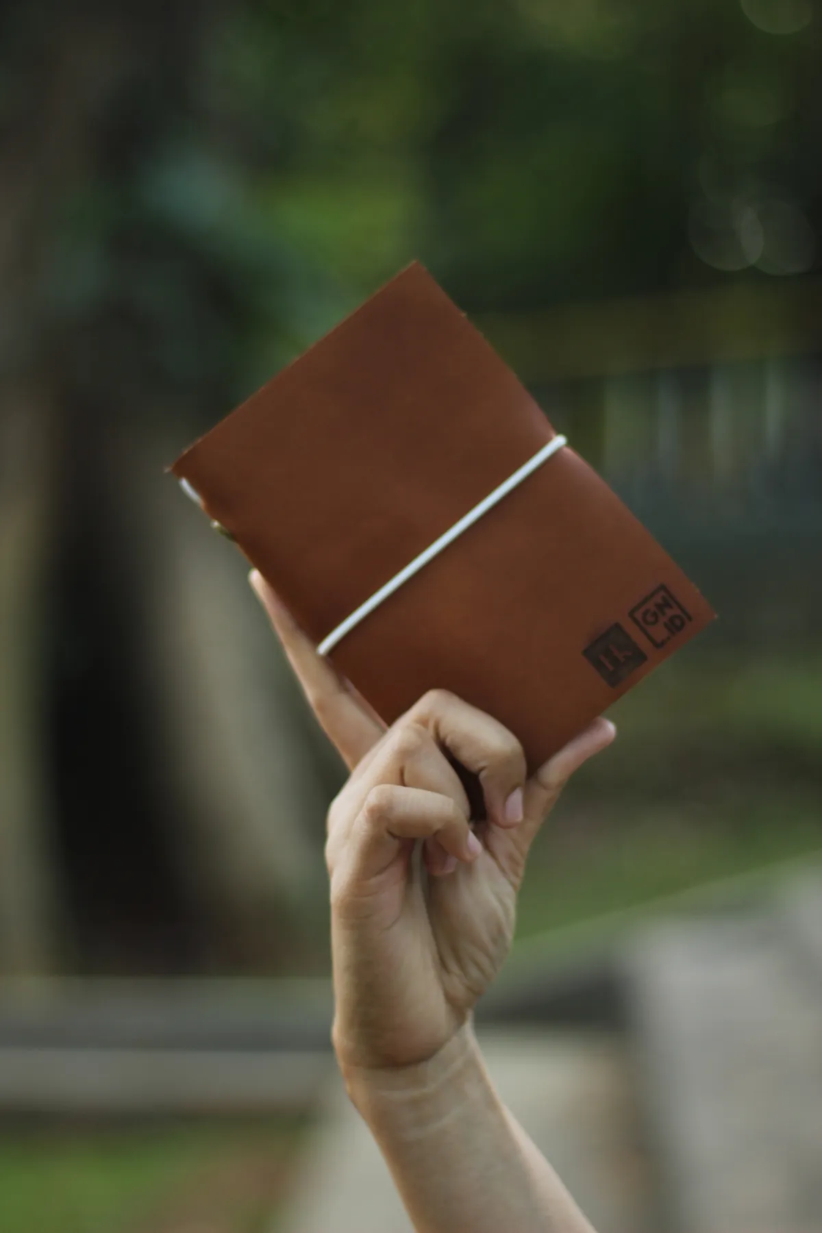A hand lifting a brown leather planner with an elastic closure, two logos debossed side by side in the lower corner of the cover