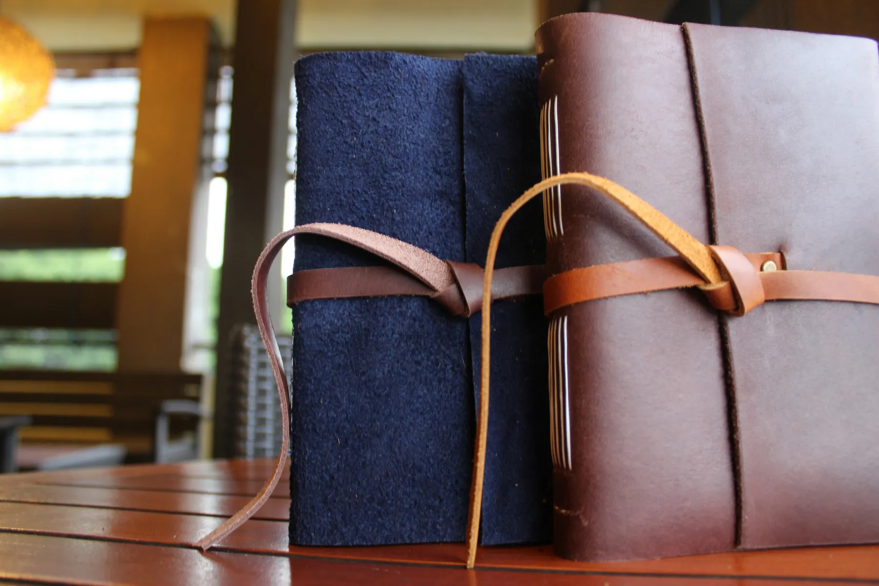 Two leather journals standing on a wooden table, one navy and one brown, each closed with a knotted leather tie