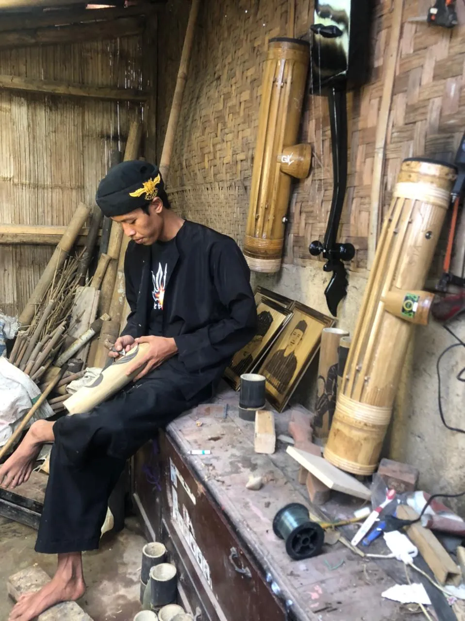 Perajin bambu duduk di bengkel beranyam bambu, meraut badan pena, dikelilingi suling dan tabung bambu