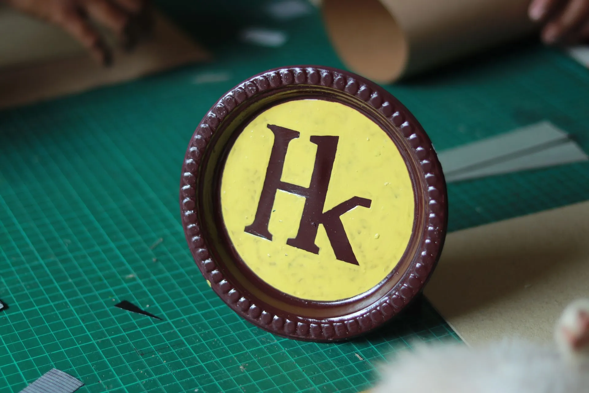Monogram Hk dicat tangan pada medali kayu bundar, di atas alas potong hijau, dengan tangan perajin bekerja di latar belakang