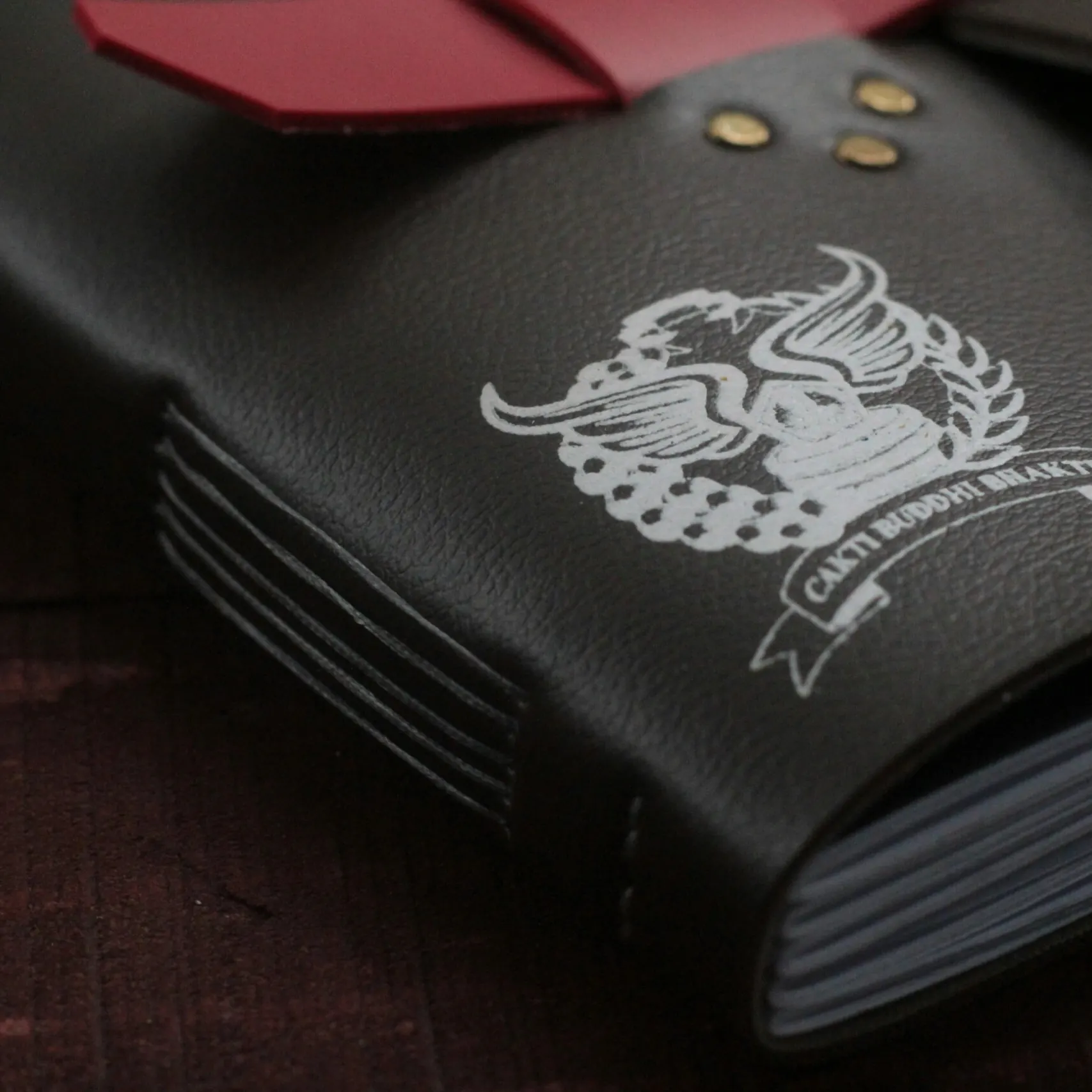 Close-up of a black leather notebook cover printed with a white Cakti Buddhi Bhakti crest, brass rivets and a stitched spine