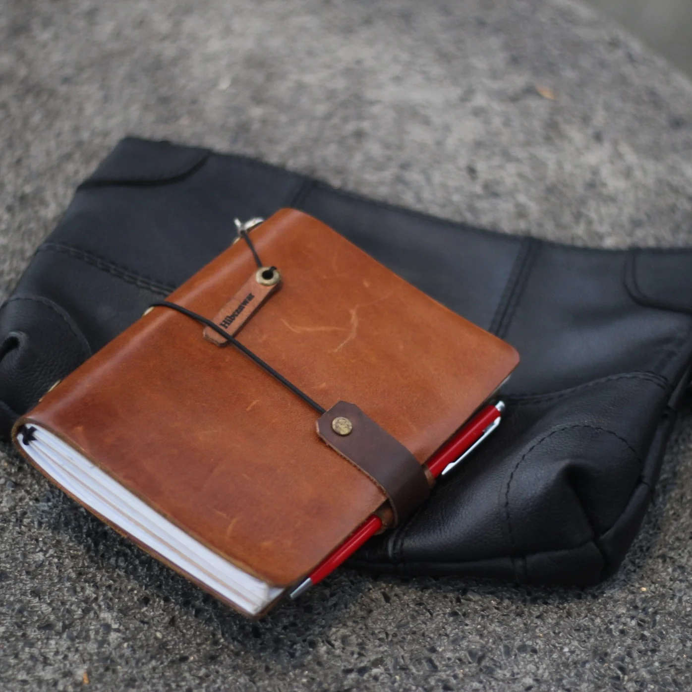 A brown leather journal with a Hibrkraft logo placed on top of a black leather pouch.