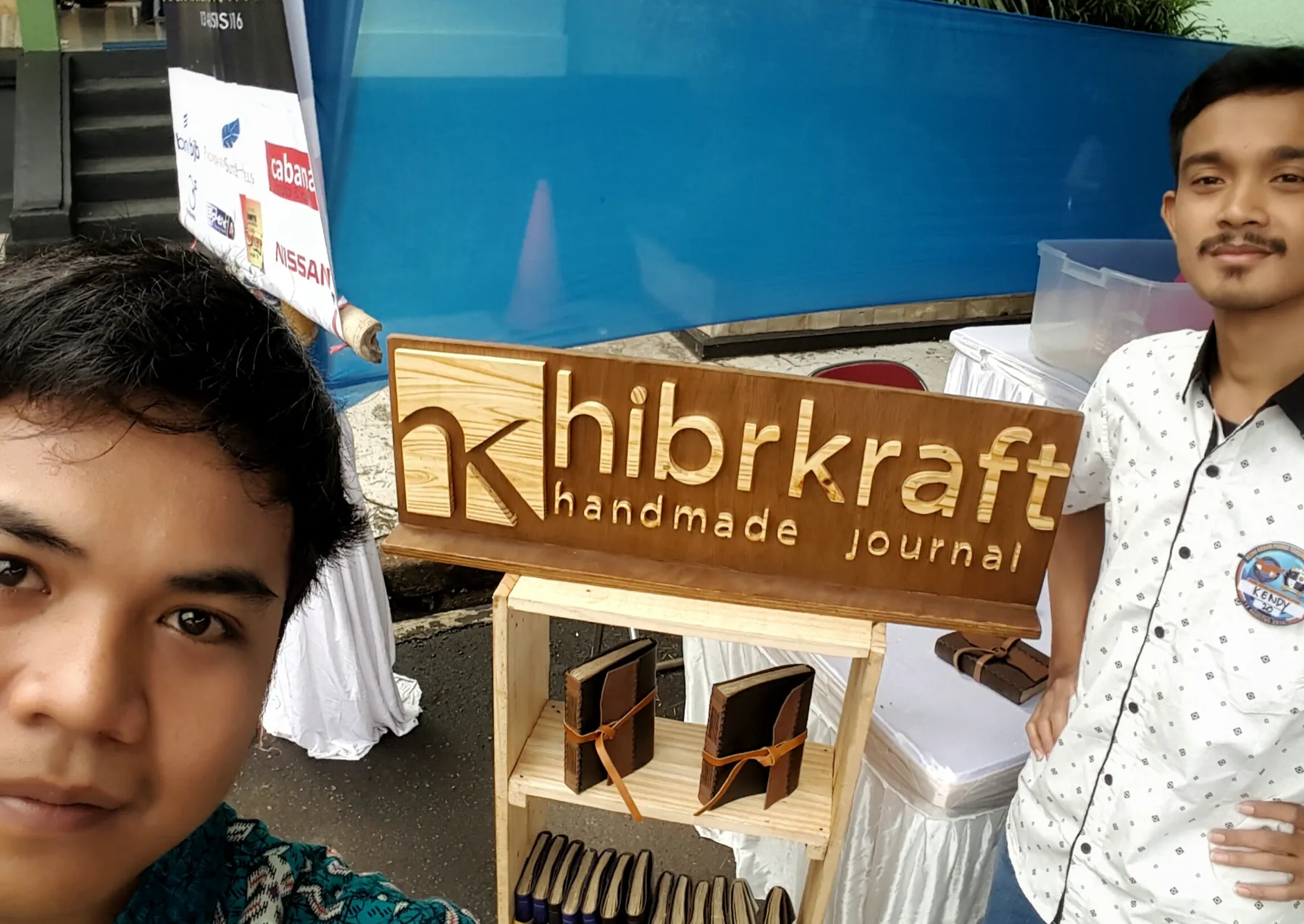 Dua pendiri Hibrkraft di booth pameran awal dengan papan nama kayu berukir dan jurnal kulit di rak