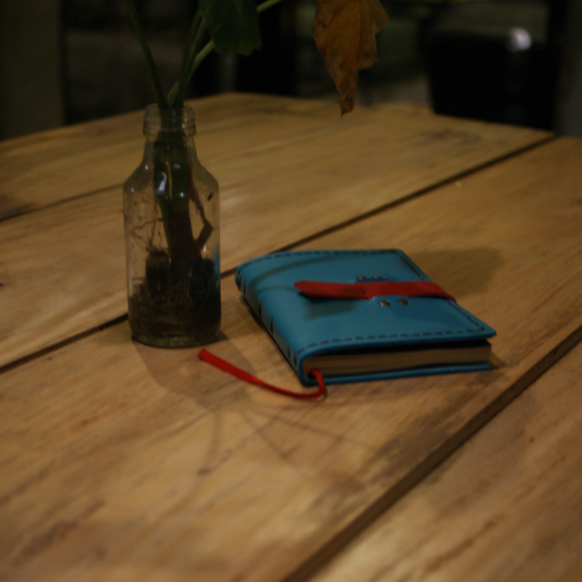 A blue leather journal with a red strap closure sits on a wooden table next to a glass vase.