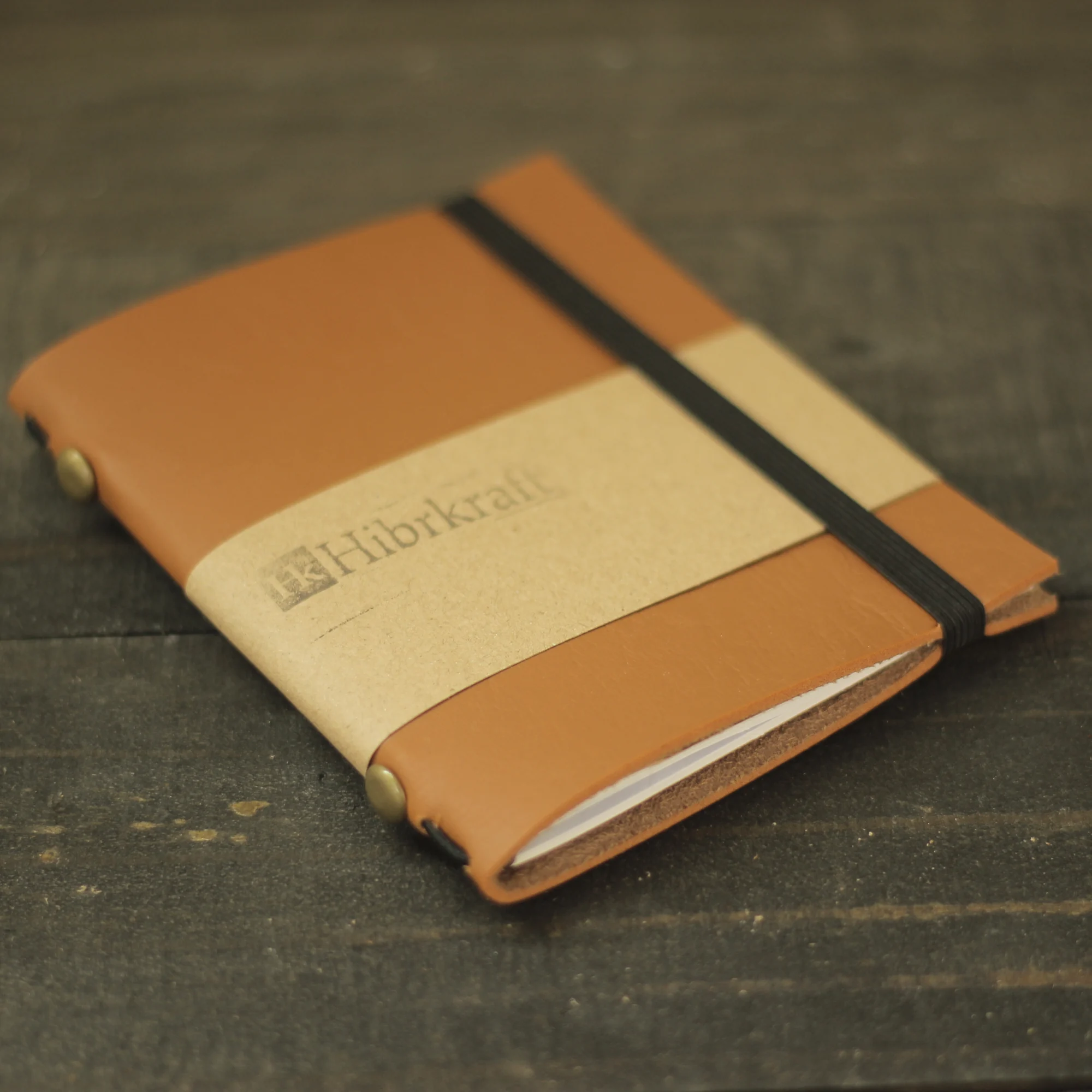 A brown leather journal with Hibrkraft branding secured by an elastic band on a wooden table.