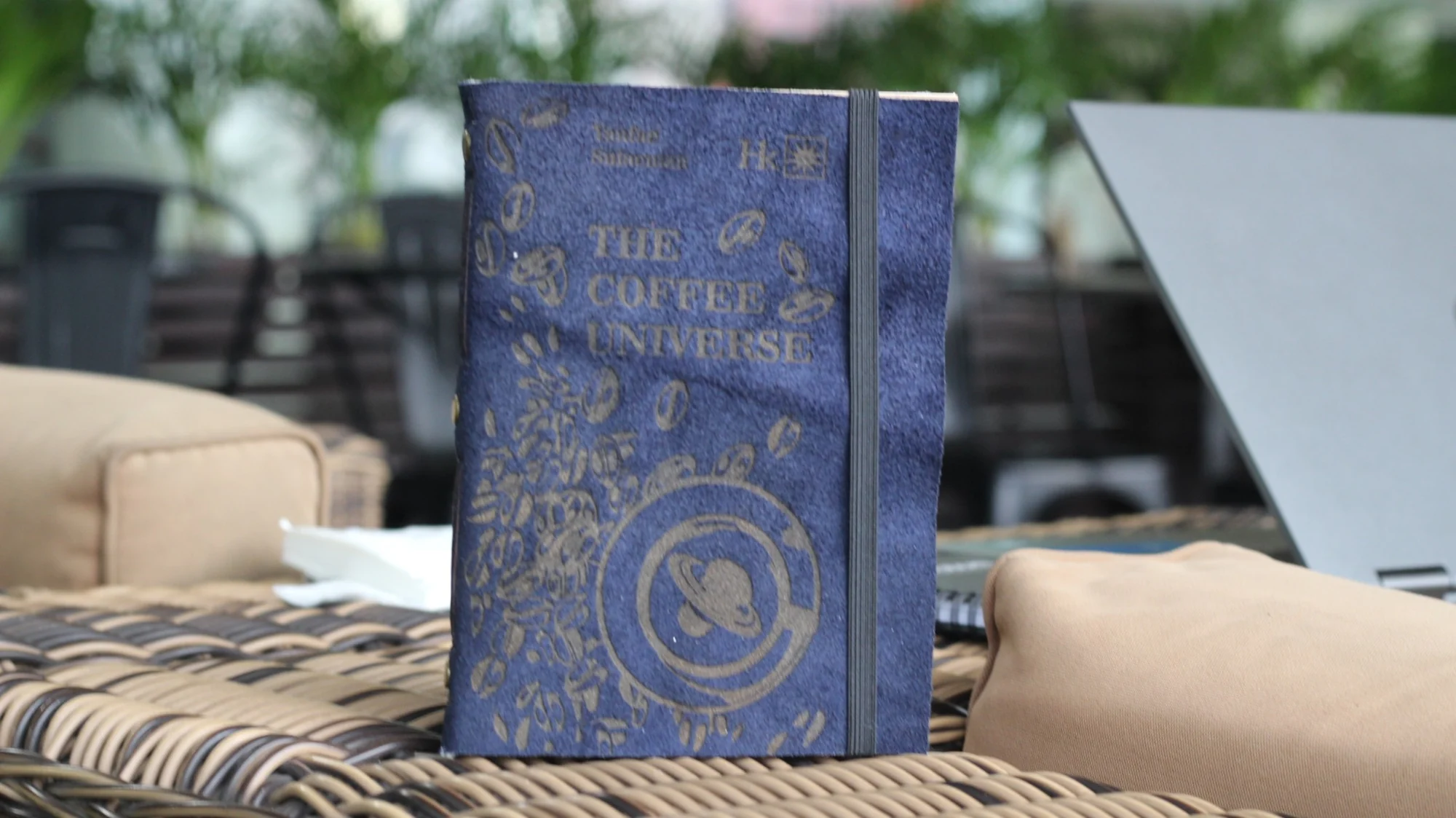Jurnal suede biru bertuliskan The Coffee Universe di atas meja anyaman