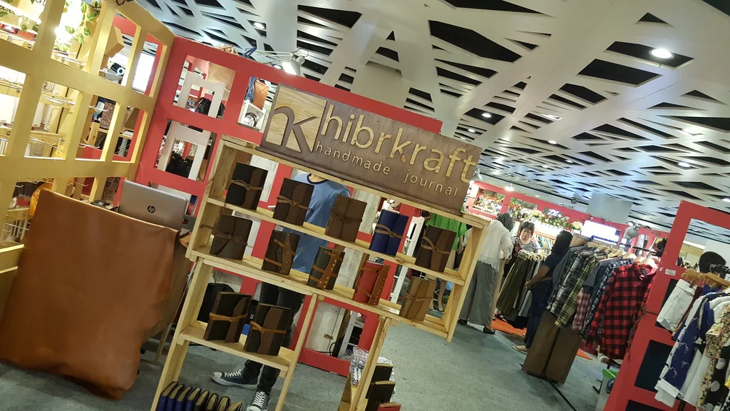 Booth pop-up Hibrkraft: rak kayu bertingkat berisi jurnal kulit bertali, papan nama kayu berukir di atasnya, sehelai kulit utuh tergelar di sisi kiri, dan lapak-lapak pakaian di latar belakang
