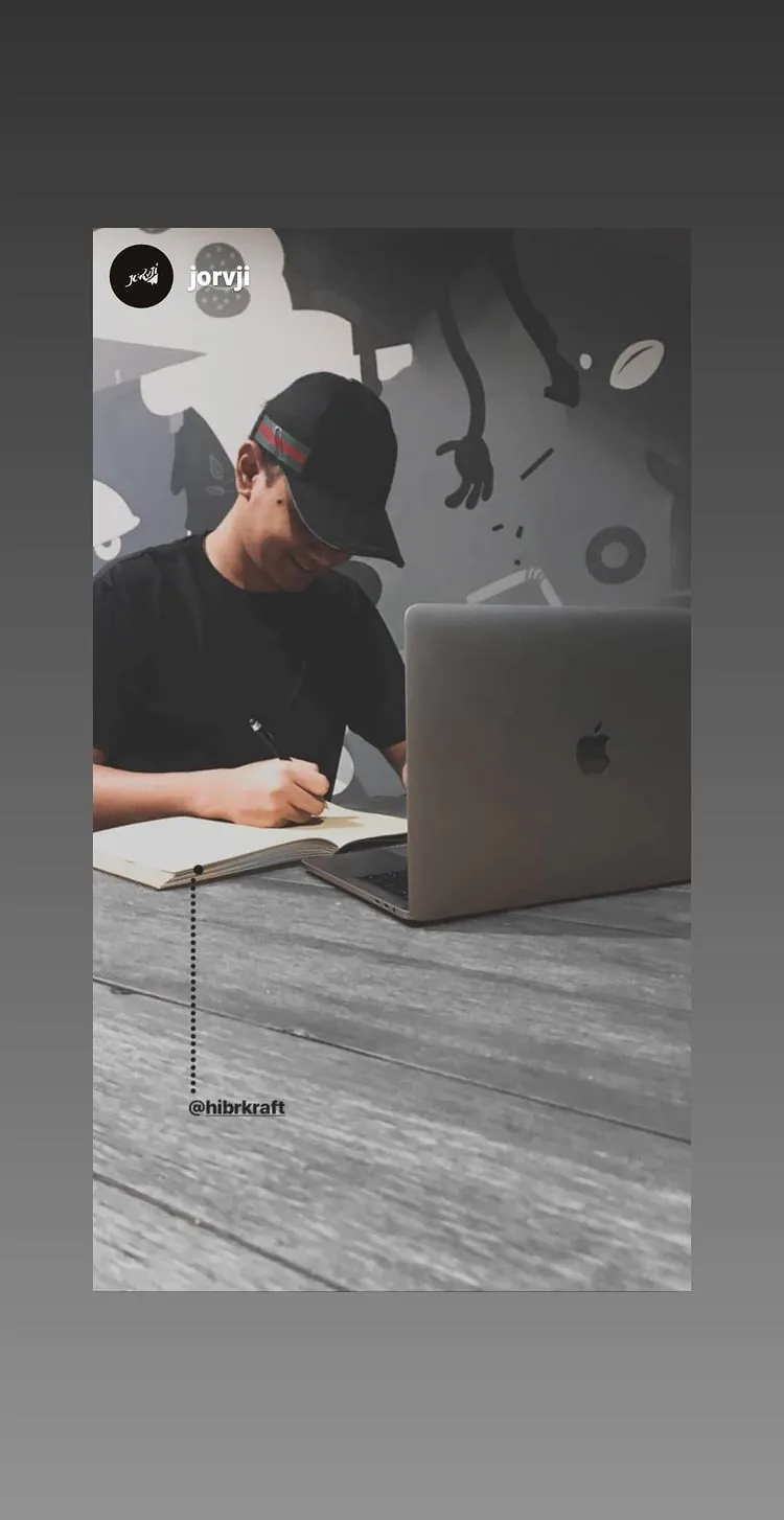 Story Instagram: pria bertopi menulis di jurnal di meja kayu di samping laptop, dinding bermural di belakangnya