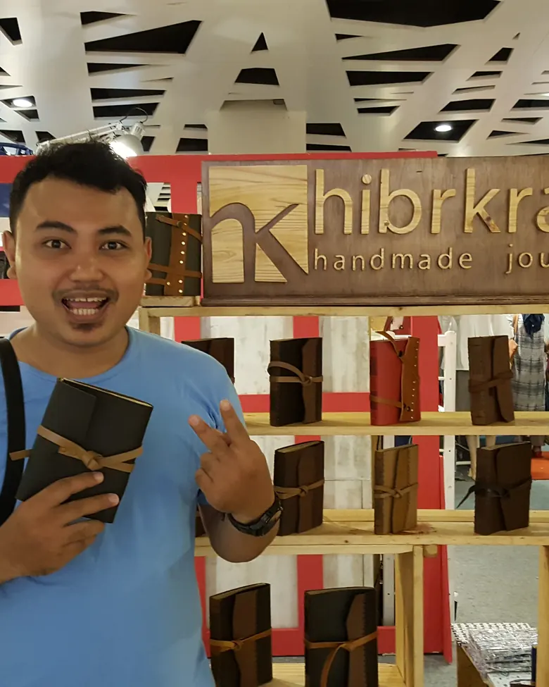 Seorang pria berkaus biru memegang satu jurnal kulit bertali di depan booth pameran Hibrkraft, dengan papan nama kayu berukir dan rak-rak berisi puluhan jurnal kulit di belakangnya