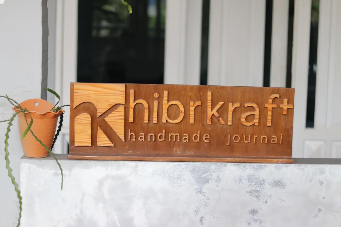 Papan nama kayu Hibrkraft dengan tanaman pot di sampingnya