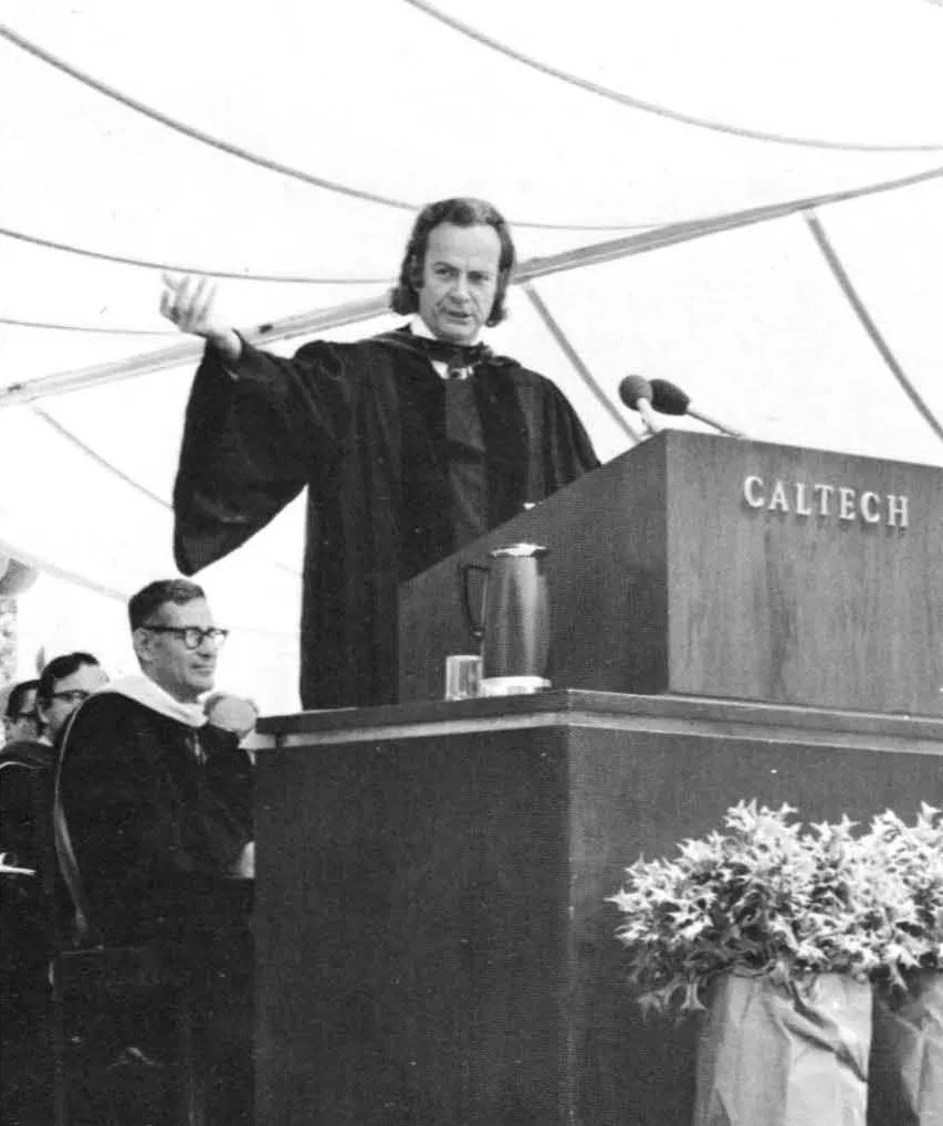 Richard Feynman mengenakan jubah akademik dan mengangkat tangan saat berbicara di podium bertuliskan Caltech, didampingi seorang kolega di sisi kiri bawah panggung