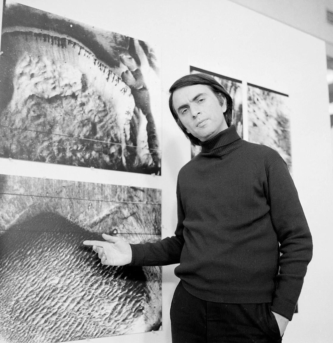 Foto hitam putih Carl Sagan mengenakan sweter turtleneck gelap, menunjuk dengan tangan ke detail pada cetakan foto permukaan Mars yang dipasang di dinding, dengan dua foto serupa lainnya terpasang di sebelah kanan setinggi kepala