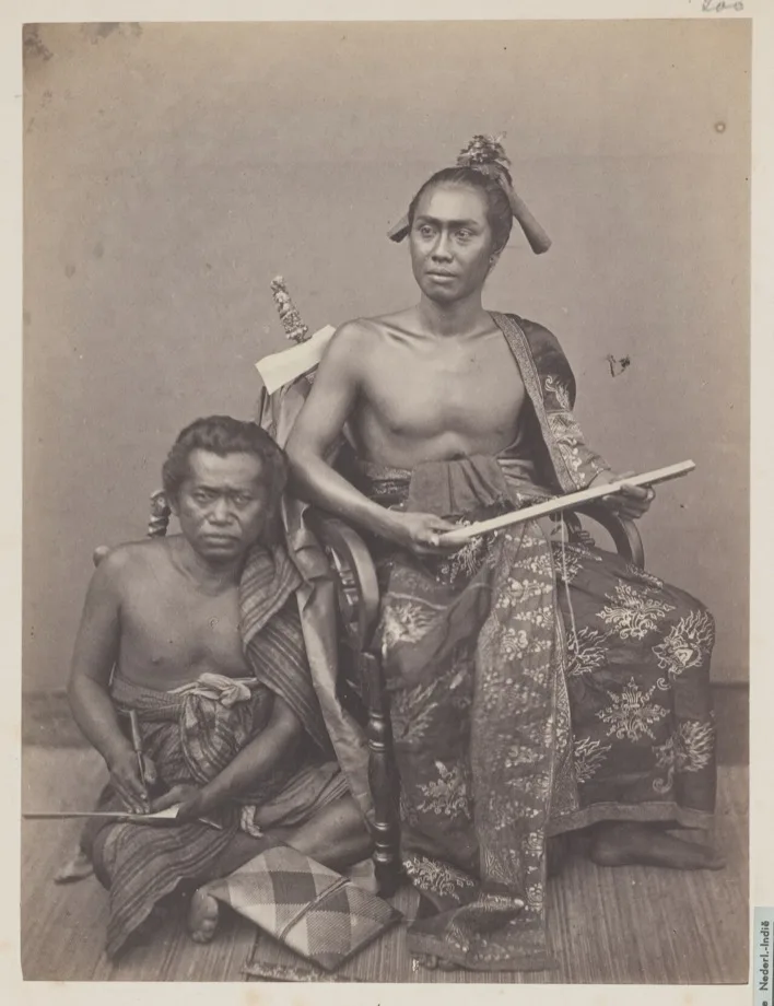Foto studio tahun 1865–1866: seorang bangsawan Bali duduk di kursi memegang lontar yang terikat tali di pangkuannya, sementara jurutulisnya duduk di kakinya sambil mengukir lembar lontar baru dengan pengutik