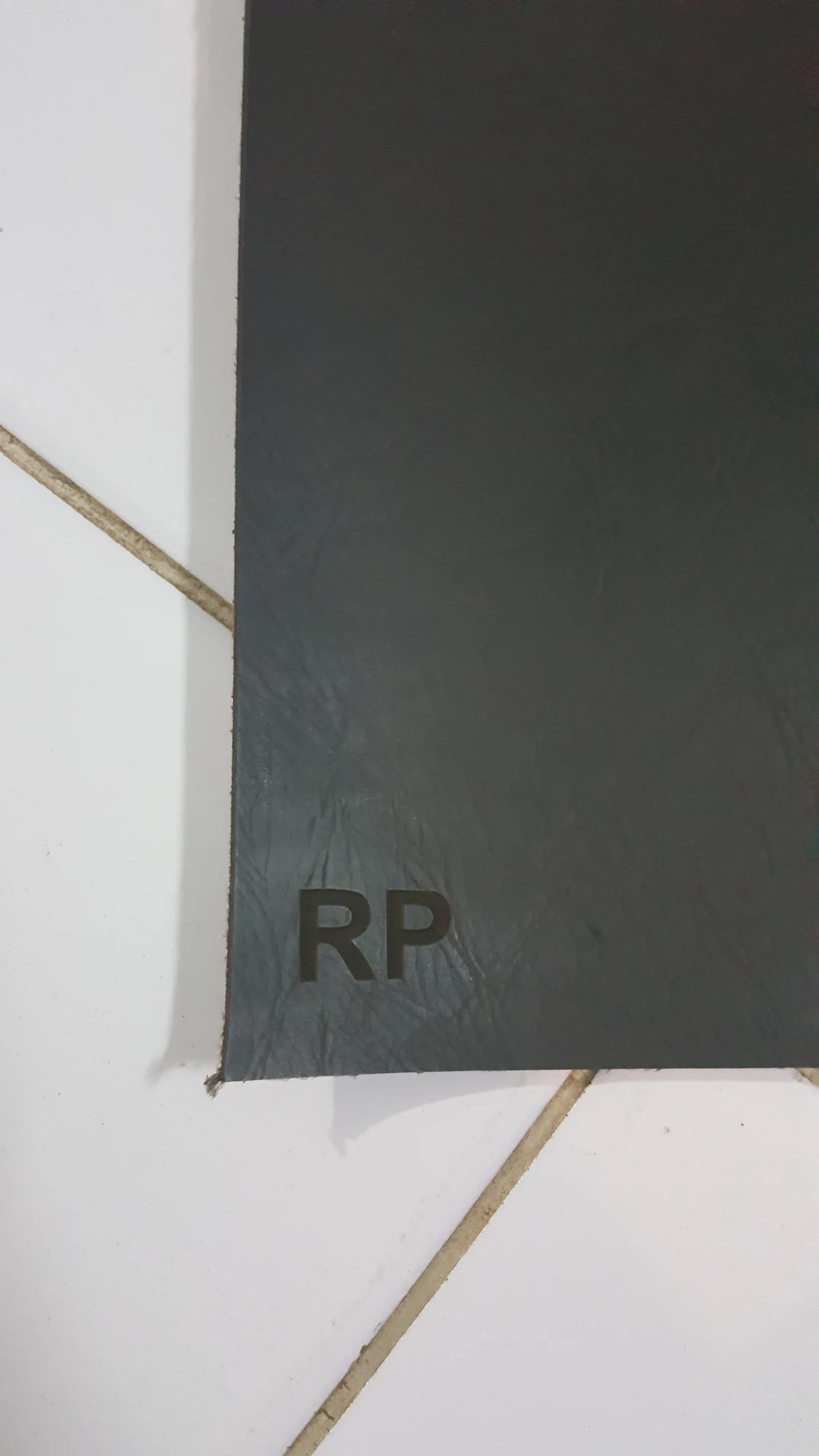 Sudut lembar kulit cokelat tua dengan inisial "RP" yang dideboss, difoto di atas lantai keramik bengkel