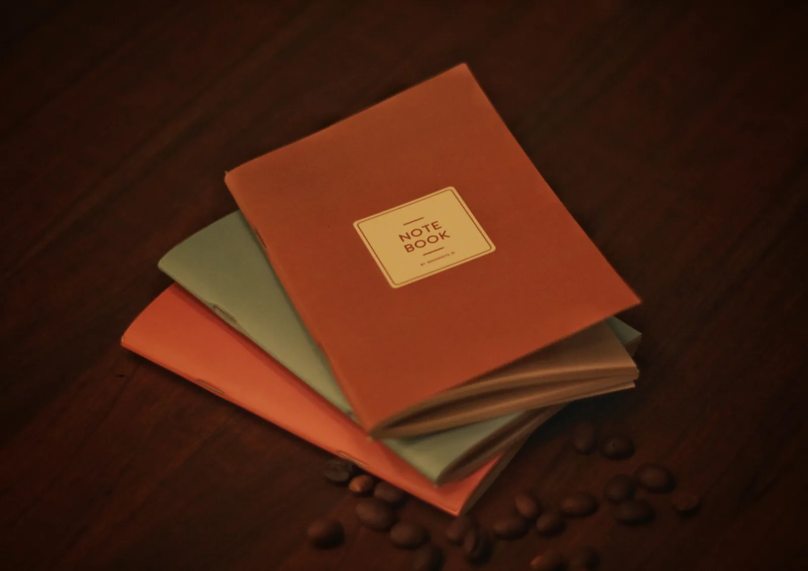 Três miolos de caderno em laranja, verde salva e castanho claro, rotulados Note Book, dispostos sobre uma mesa de madeira escura com grãos de café