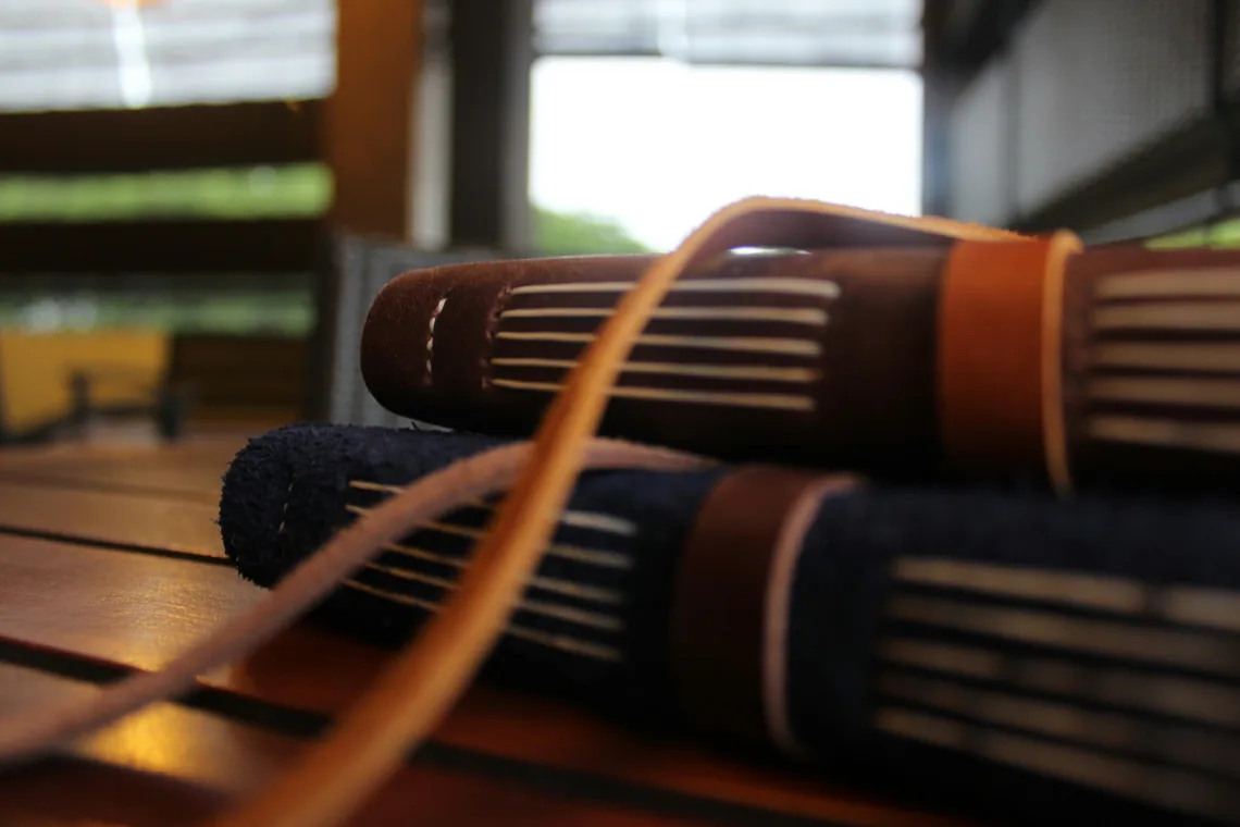 Two journals stacked on a slatted wooden table, spines toward the camera, one dark navy and one brown, a long tan leather tie trailing across them