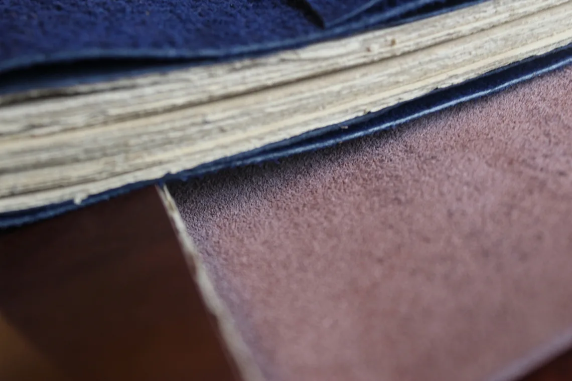 Macro view of a journal's fore-edge, showing the cream page block in cross-section between a dark navy suede cover above and a brown leather surface below