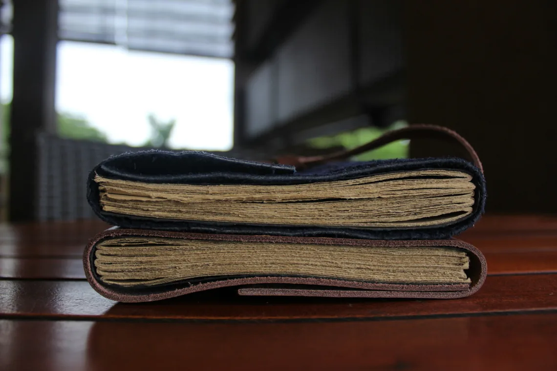 Two journals stacked side-on on a dark table, their thick text blocks of irregular-edged pages clearly visible, the upper navy and the lower brown