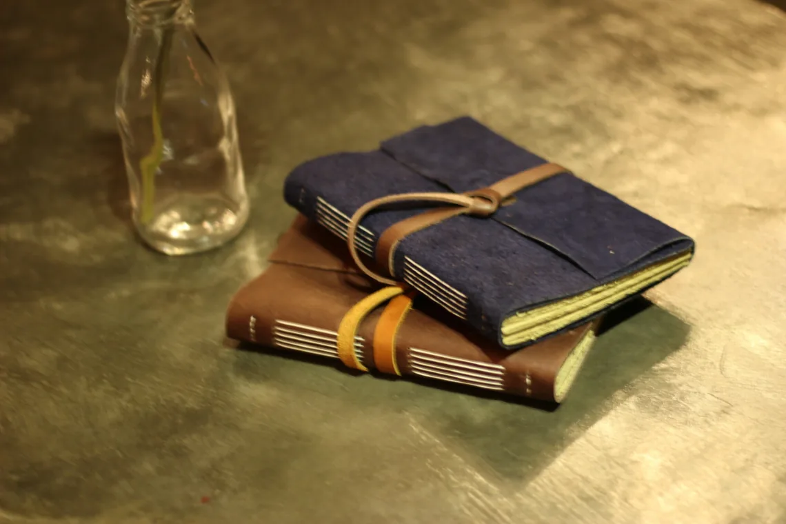 Two coptic-bound journals stacked beside a clear glass bottle on a brass surface, the upper one navy with a tan tie, the lower brown with a yellow tie