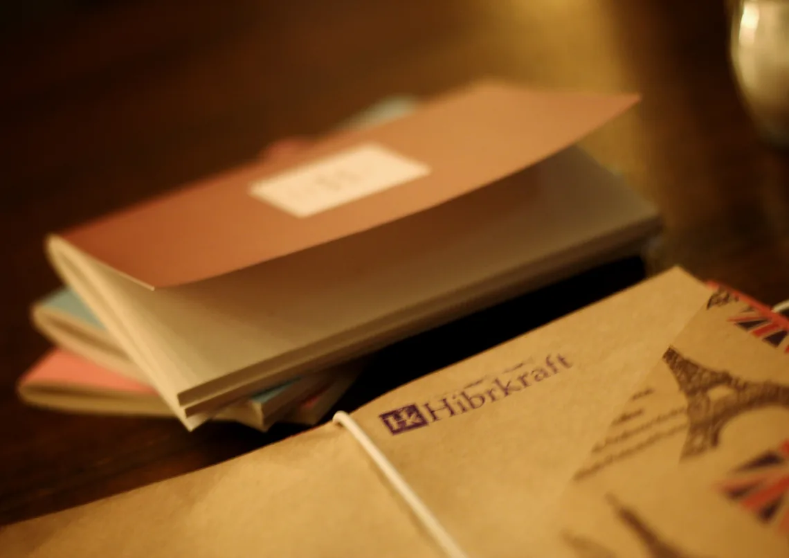 A fanned stack of cream and pastel note cards in front of a kraft paper sleeve printed with a travel motif, beside a small booklet closed with a white elastic and carrying a Hibrkraft label